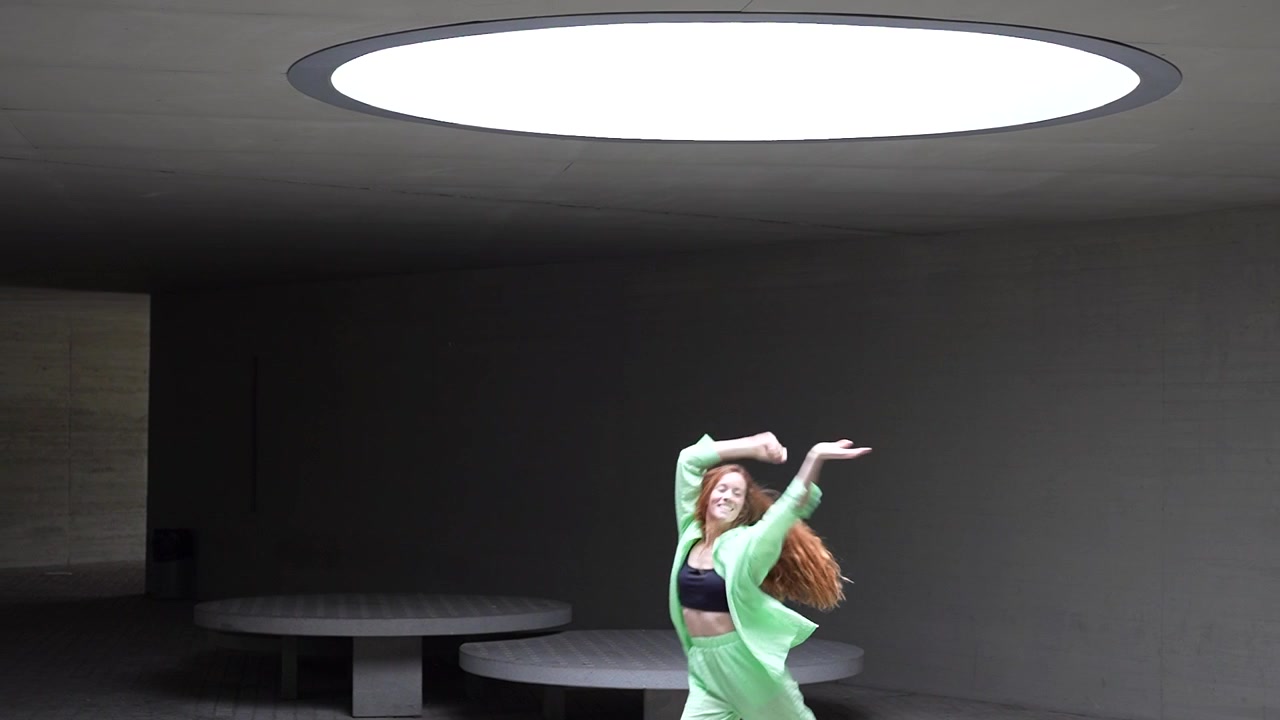 Una ballerina con un abito al neon esegue mosse dinamiche sotto un lucernario con una coreografia accattivante.