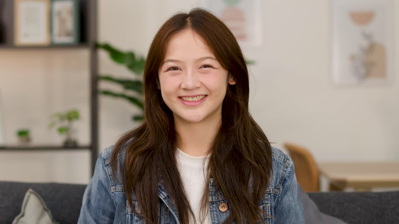 หญิงสาวร่าเริงยิ้มอย่างมั่นใจบนโซฟาในร่ม สวมเสื้อแจ็กเก็ตเดนิมลำลอง ภาพถ่ายระยะกลางที่มั่นคง