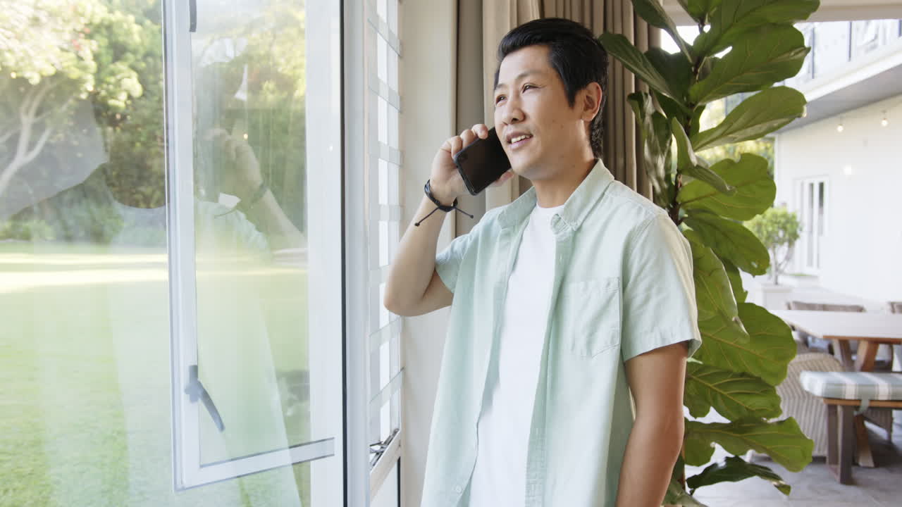 Talking on smartphone, asian man smiling and looking out window in bright room