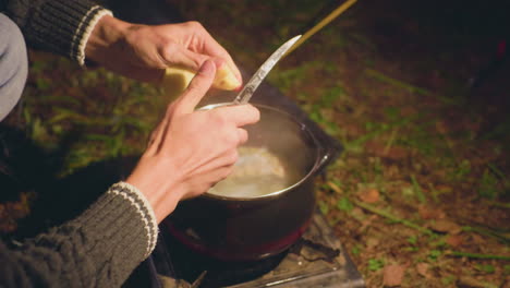 man peels potatoes directly over steaming pot during night camping in forest, seated near ground with portable stove on grassy terrain, focused on meal preparation under soft warm light