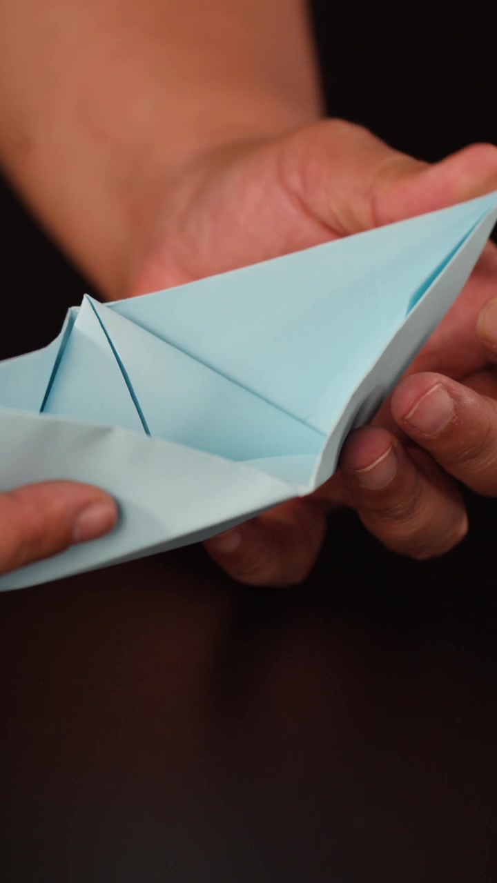 Primo piano di mani che piegano con cura carta origami celeste su un tavolo scuro, con luce soffusa da studio.