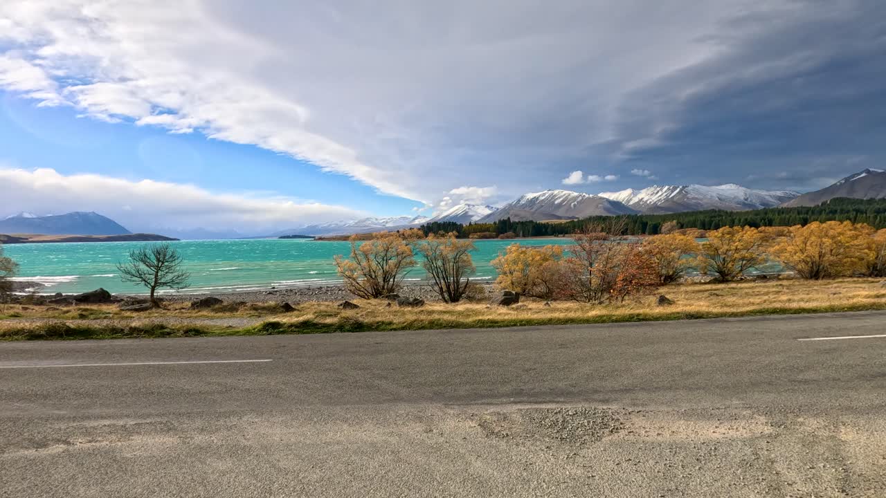 A vehicle approaches a vibrant turquoise lake surrounded by autumn foliage, rugged mountains, and dramatic clouds, with dynamic camera movement and natural daylight