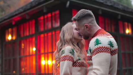 romantic couple standing hugging near the country house with red lights inside. young people dating and kissing outdoors.
