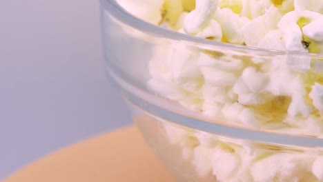 Extreme-close-up-of-popcorn-in-transparent-glass-bowl-rotating-on-orange-surface,-macro-shot-view-in-4k