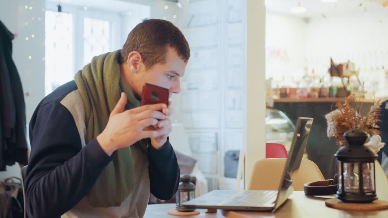 Uomo bianco che digita al laptop in un caffè, studente concentrato che alterna ricerca e presa di appunti, controllo occasionale del telefono, sorseggiando caffè da una tazza, indossa una sciarpa verde, luce calda naturale dalla finestra.