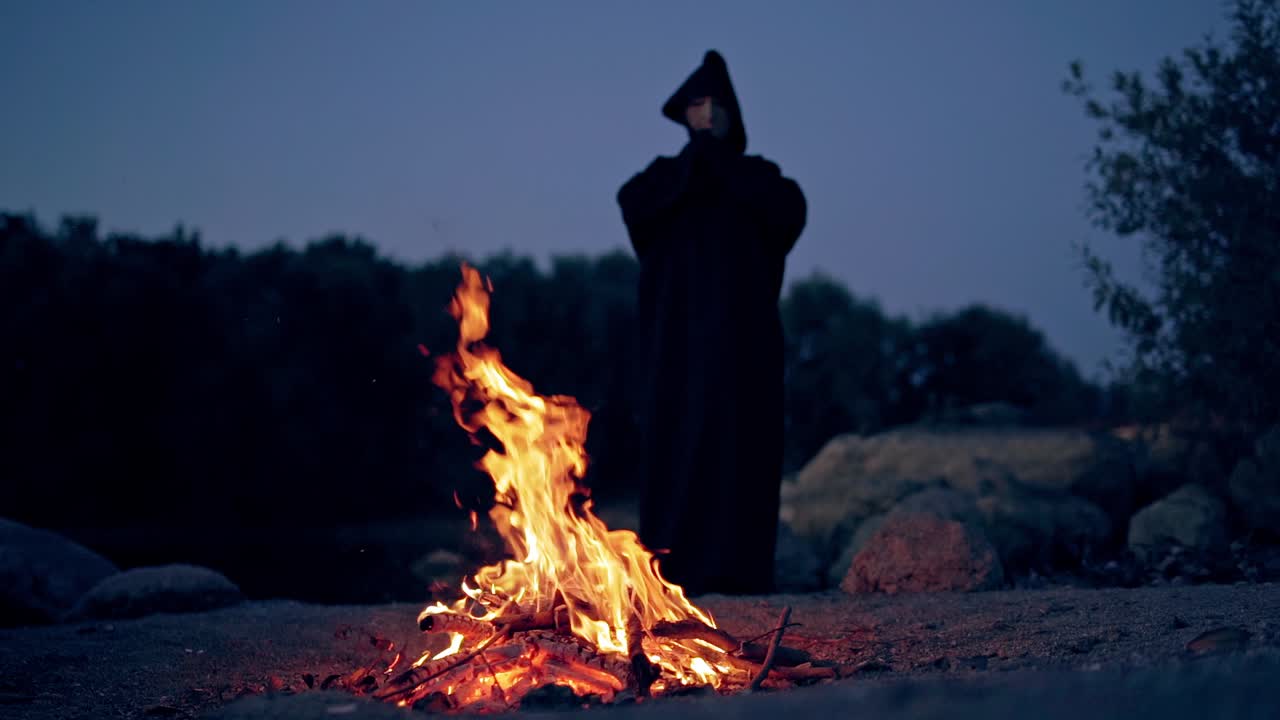 Halloween. Powerful witch or wizard in black costume stands and watches the fire flame in the evening.