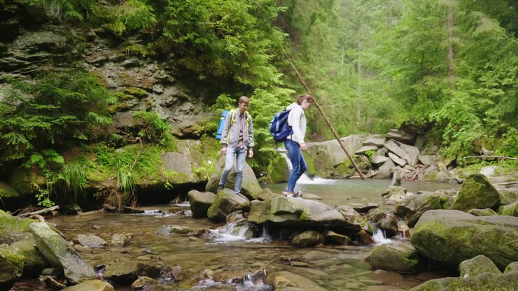 Una giovane coppia di turisti con zaini attraversa un fiume di montagna nella foresta. Video HD.