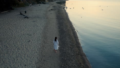 Filmische-Drohnenaufnahme-Einer-Schönen-Frau-In-Weißem-Kleid,-Die-Allein-Am-Einsamen-Strand-Spaziert