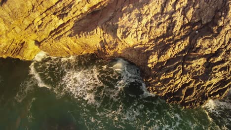 waves crashing cliffs on sunny evening, aerial downwards view