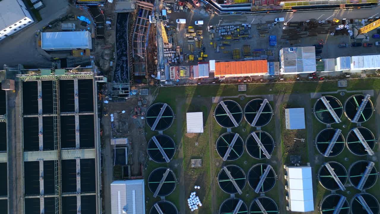 Industrial complex with a chimney emitting smoke into the sky, next to a sewage treatment facility and urban infrastructure. Unique aerial view overflight flyover drone