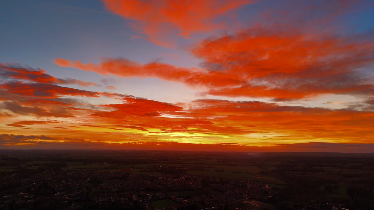 Aerial drone view of dramatic sunset with vivid orange red clouds over wide landscape and distant town creating colorful sky and calm evening atmosphere