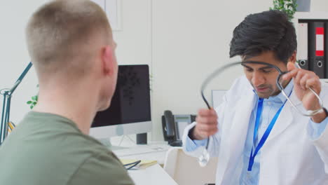 male doctor or gp wearing white coat examining young man listening to chest with stethoscope