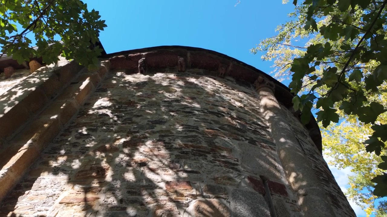 Low-angle view of the curved stone exterior of San Miguel de Xagoaza, highlighting weathered masonry and historic religious architecture beneath leafy trees in Galicia