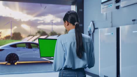 back view of asian female with home energy storage system in a modern garage, typing on laptop's keyboard with green screen and look at the factory once