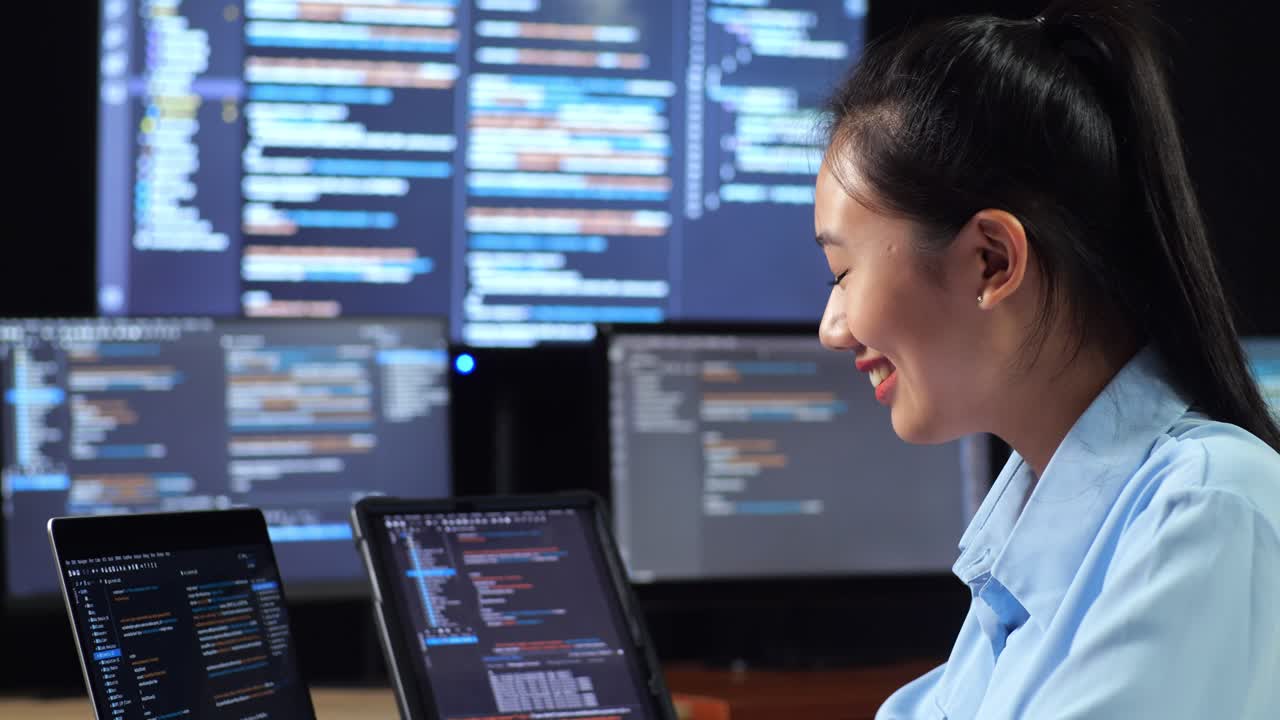 vista lateral de cerca de una programadora asiática que mira la base de datos en una tableta y escribe código en una computadora portátil utilizando múltiples monitores que muestran la base de dados en los escritorios de la oficina