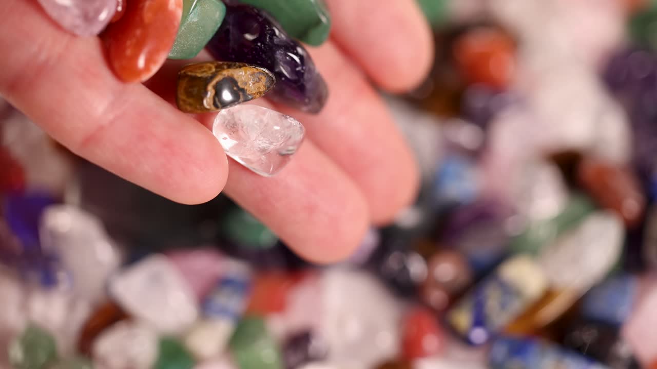 A hand releases vibrant chakra stones onto a surface, showcasing their colors and textures in soft lighting