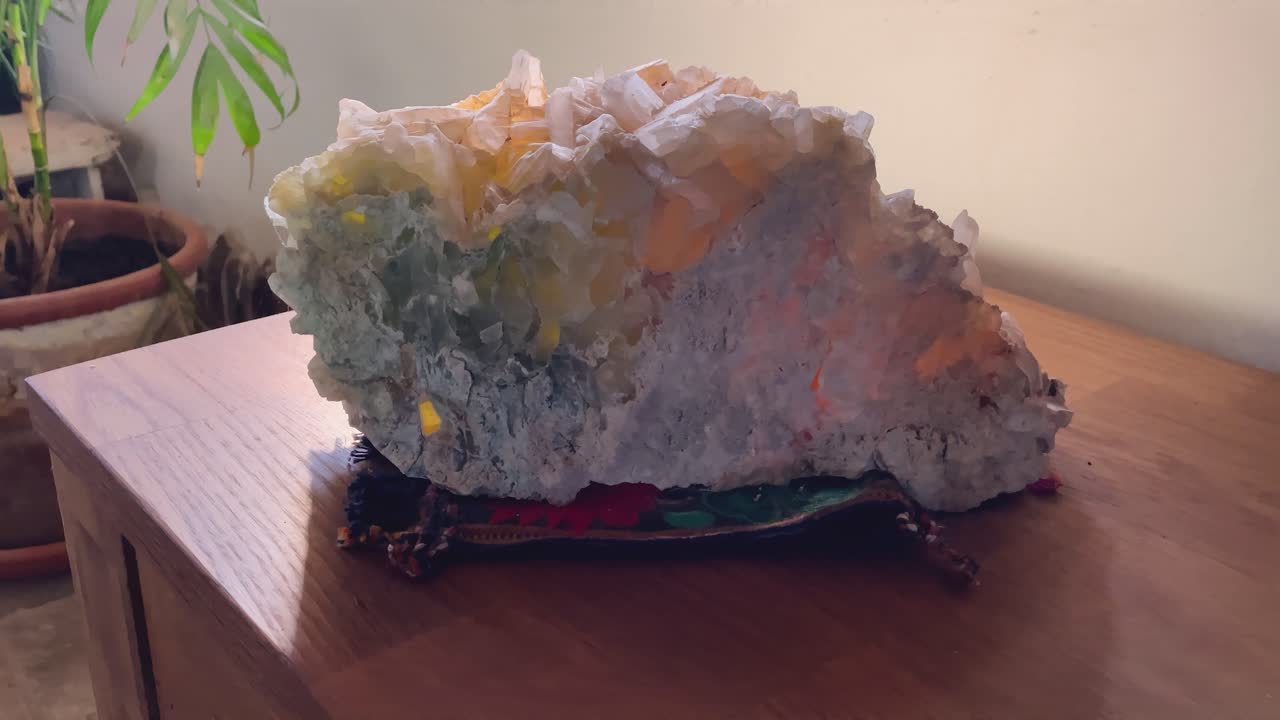 Filmagem em câmera lenta de um cristal grande de lapis specularis com tons esverdeados e alaranjados. O mineral repousa sobre um tecido decorativo em uma superfície de madeira, parcialmente iluminado pela luz suave da janela.