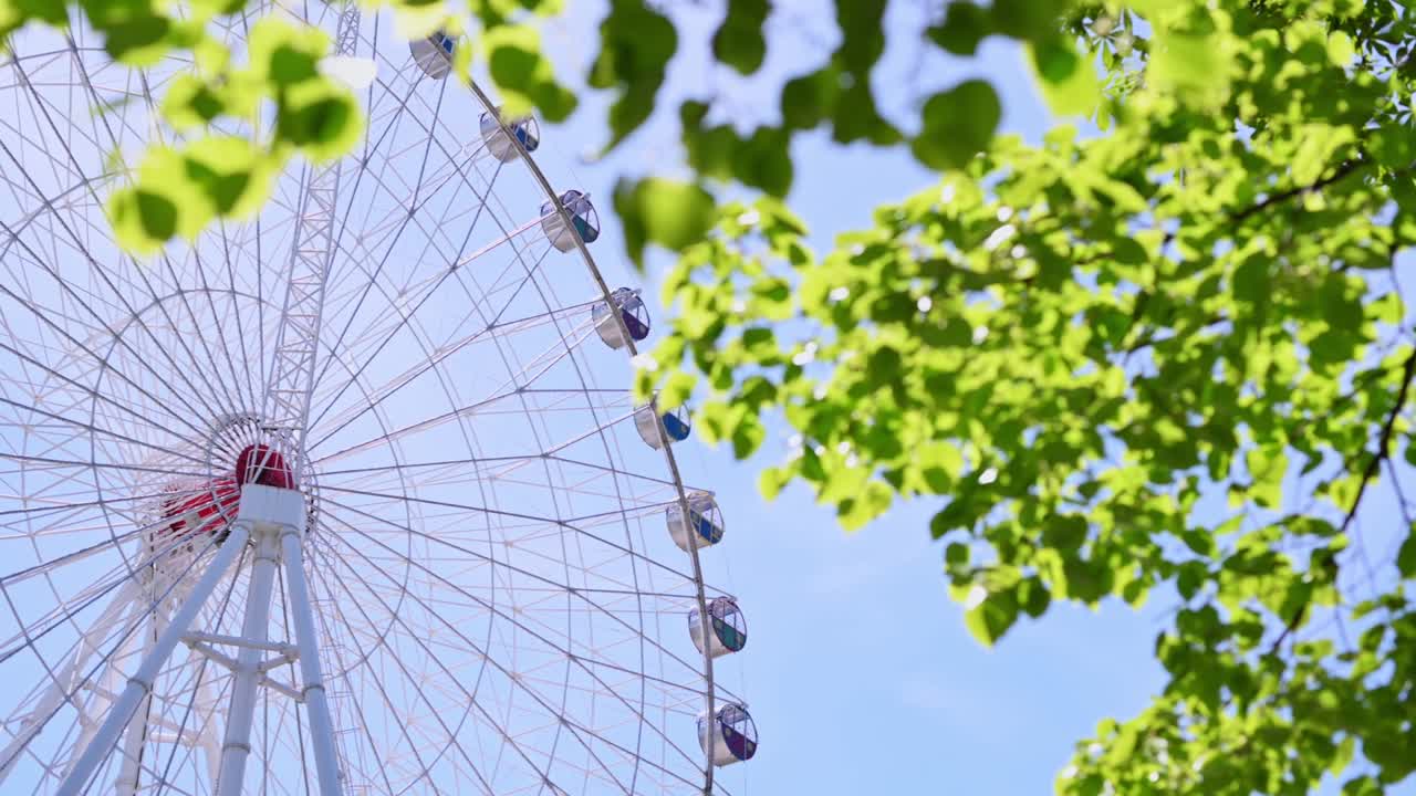 다채로운 오두막을 가진 페리스 의 전망, 공원의 봄 녹색 나무 잎을 통해, 파란 하늘에 대한 도시 페리스 바, 엔터테인먼트 카루셀, 축제, 레크리에이션, 재미있는 활동