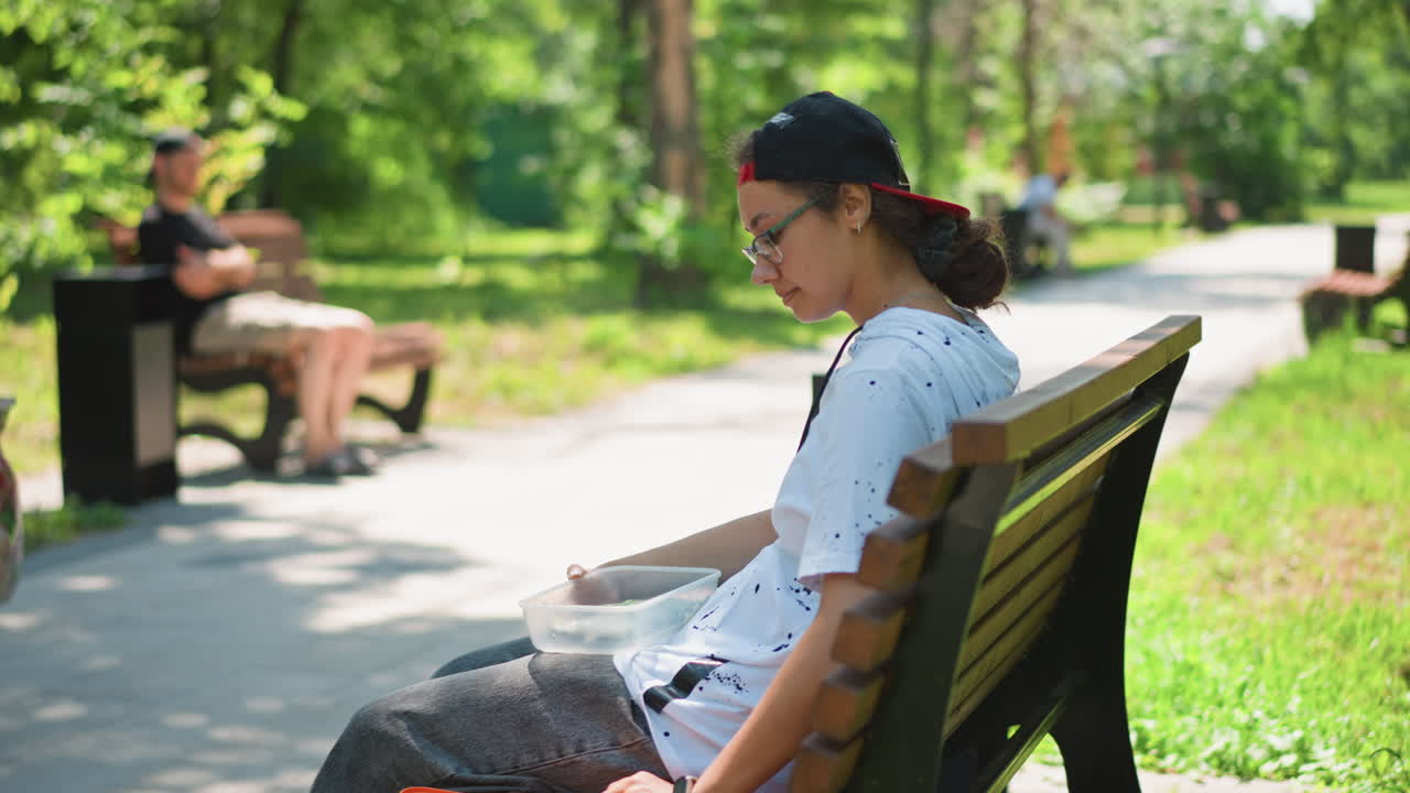 Individual arranges food box next to trash, Person sets down food holder beside park trash, An individual discards food container while observing surrounding park scenery and pedestrians