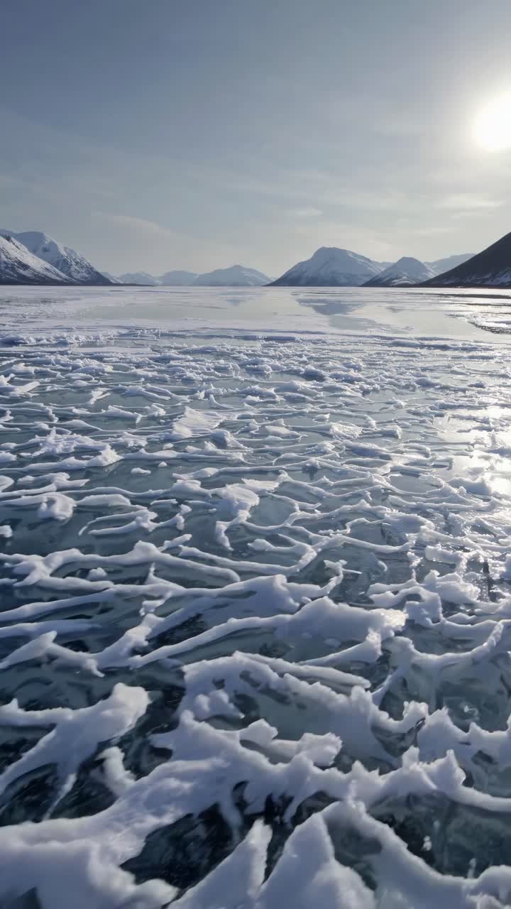 Aerial video captures a frozen lake with intricate ice patterns, surrounded by snow-capped mountains