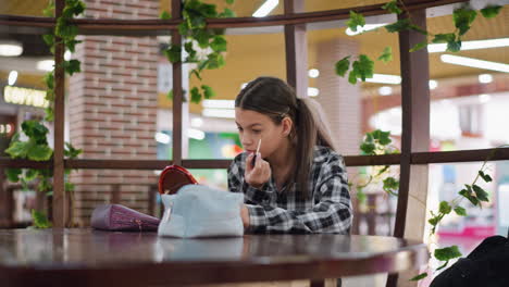 dama sentada en la mesa del café aplicando maquillaje usando un pequeño espejo y un pincel cosmético, expresión enfocada mientras mezcla cuidadosamente la fundación, ambiente acogedor con plantas crea un entorno relajado