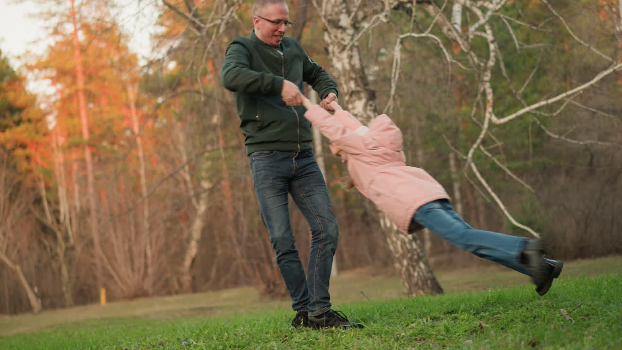 A joyful moment captured as a man in a green jacket and blue jeans spins a little girl in a pink jacket around on a lush green field in a park. The scene embodies happiness, family bonding