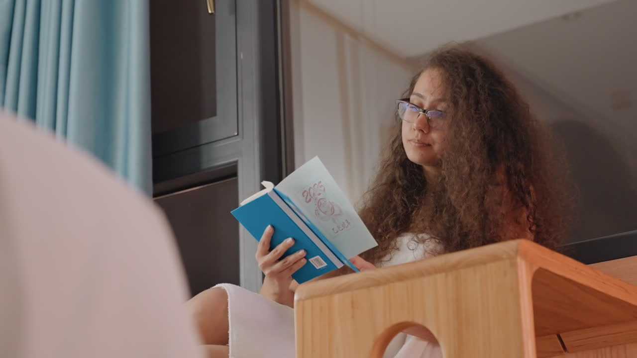 Femme lisant près de la fenêtre d'un hôtel en gros plan, lunettes encadrant un regard pensif, livre bleu posé sur une table de chevet en bois, douce lueur de lampe et rideaux en arrière-plan, étudiante ou écrivaine concentrée révisant des pages, calme