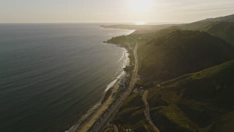 aerial drone shot flies right from a malibu sunset over pacific coast highway pch to a green mountain at golden hour