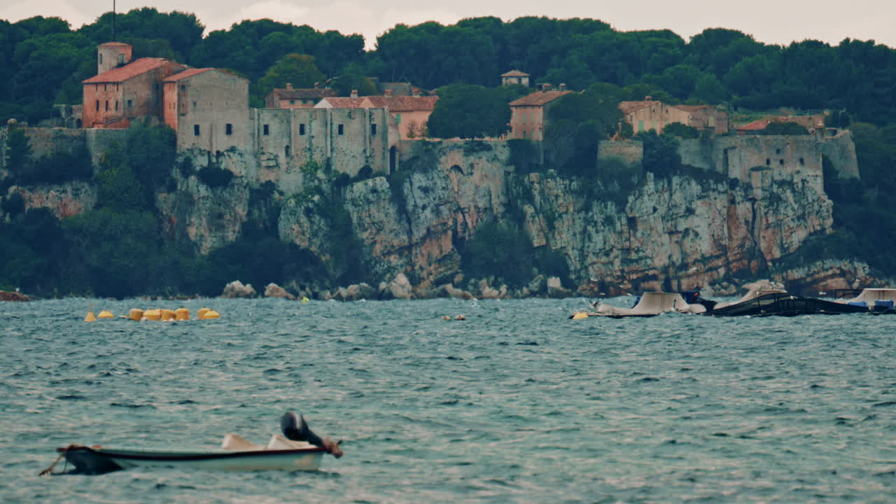 A medieval stone fortress and village perched on a high cliff above the Mediterranean Sea, with a fish farm and moored platforms spread across the water below