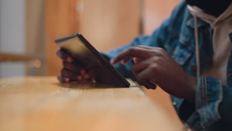 close up black hand holding tablet at angle on wooden table while finger taps screen indoors with denim jacket sleeve and hoodie detail soft bokeh warm ambient background