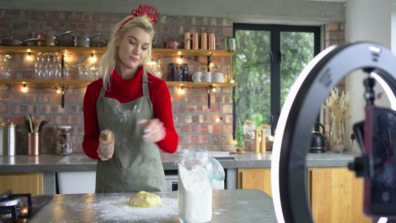 Donna che indossa un grembiule che nota un avviso sul telefono, mentre prende la farina, impasta e stende la pasta per una torta in cucina
