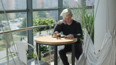 young man with blond hair enjoying a sandwich and iced coffee at a cafe table by a window with city views, natural light, and potted plants. relaxed and casual dining scene