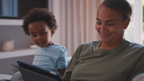 mother with son playing game or streaming to digital tablet sitting on sofa at home together