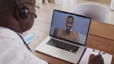 african american male doctor wearing phone headset taking notes while having a video call on laptop