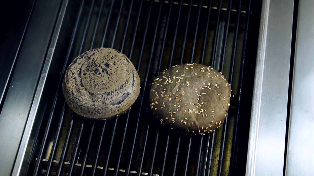 Frying buns on kitchen grill. Chef's hand putting halves of black buns for cooking burger on hot grill. Grain bread for hamburgers is grilled