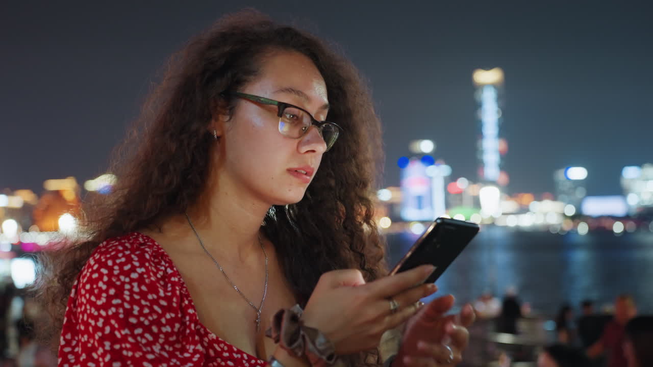 Donna sul lungomare di notte che scrive al telefono, giovane donna in vestito rosso che scorre i messaggi con lo skyline della città sfocato sullo sfondo, bagliore dello schermo che si riflette sugli occhiali, turisti e luci fuori fuoco, contemplativa.