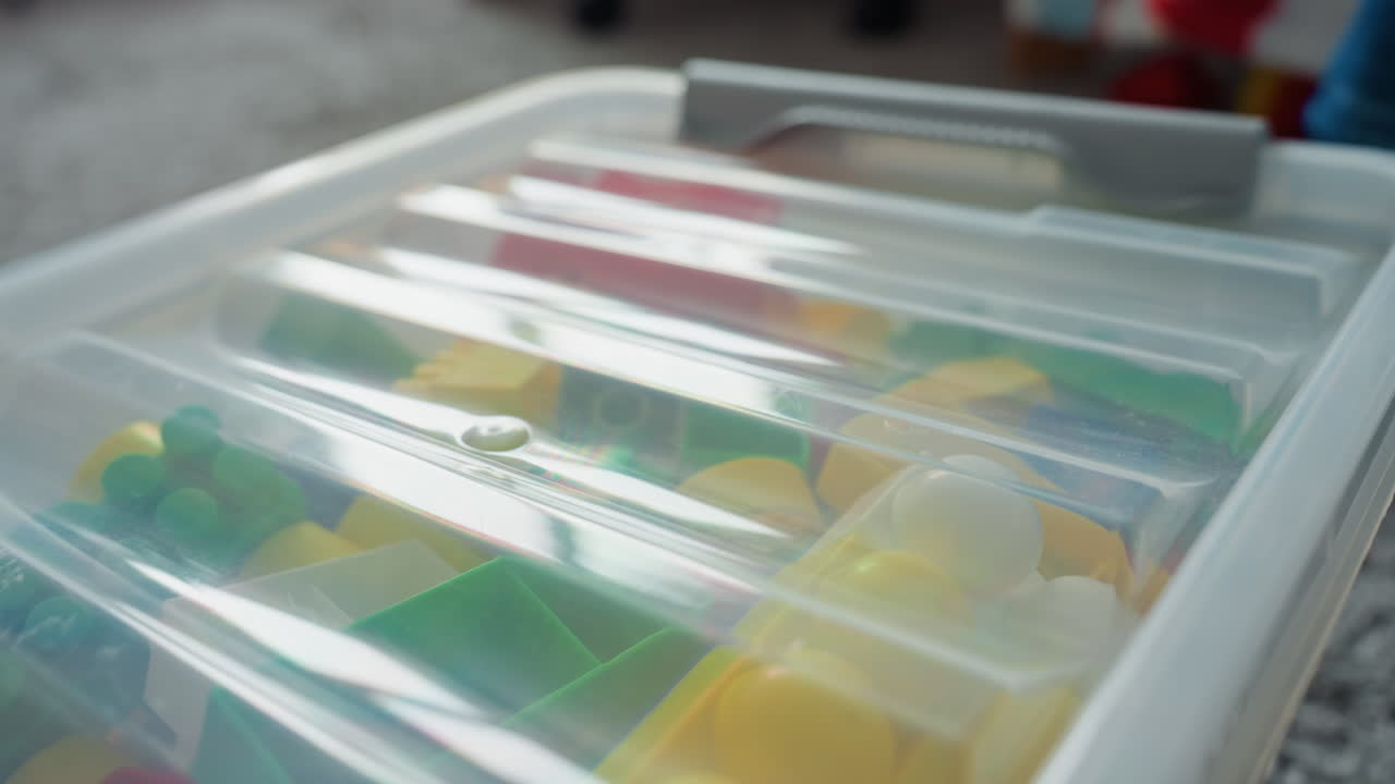 Gros plan sur un couvercle de coffre à jouets en plastique, couvercle translucide et loquet sécurisé révélant des blocs colorés flous à l'intérieur, fond de tapis doux, détail de rangement soigné pour l'organisation de la salle de jeux et la texture du produit.