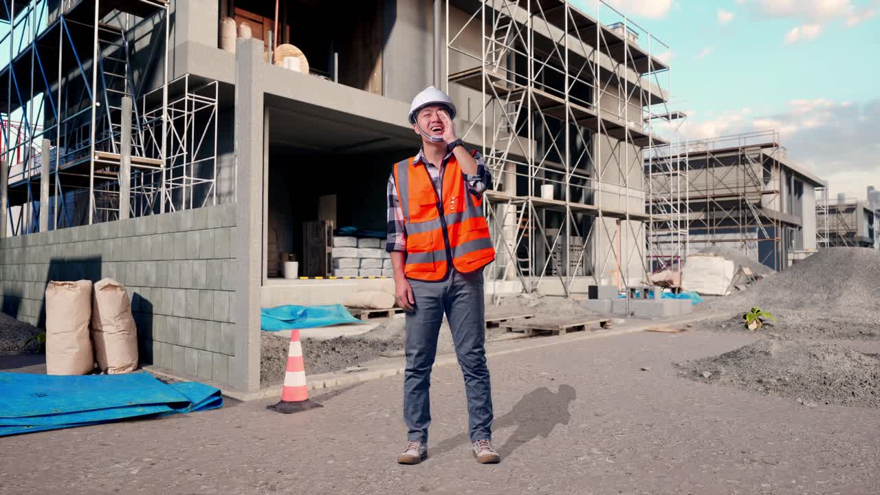 Full Body Of Asian Male Engineer With Safety Helmet Yelling With Hand Over Mouth While Standing At Building & Construction Site in Progress to New House