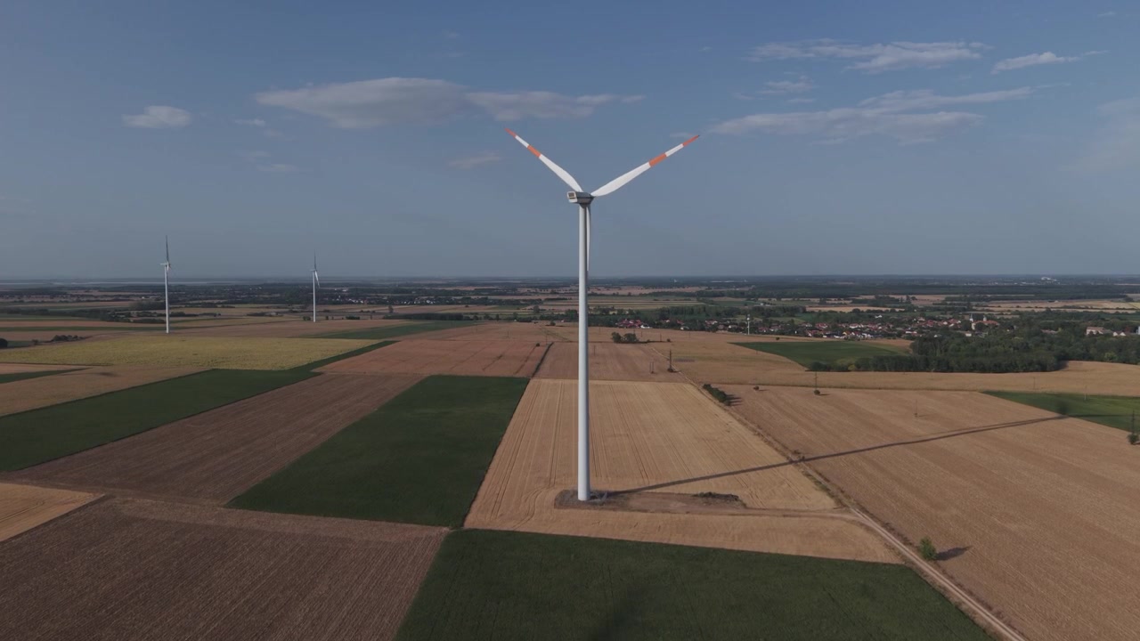 Aerial view of a large wind turbine standing among expansive agricultural fields, with additional turbines and a rural village visible in the distance.