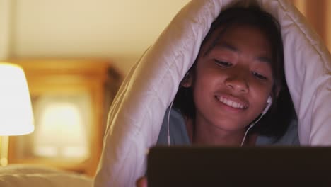 la chica asiática está usando la tableta para escuchar música y cantar de acuerdo con el ritmo en la cama con auriculares en la noche