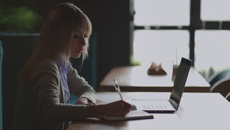 asian woman working on her laptop and writing in his notebook sitting at a table. working in coffee shop. woman looking to a laptop screen and making notes in her notebook. studying online.