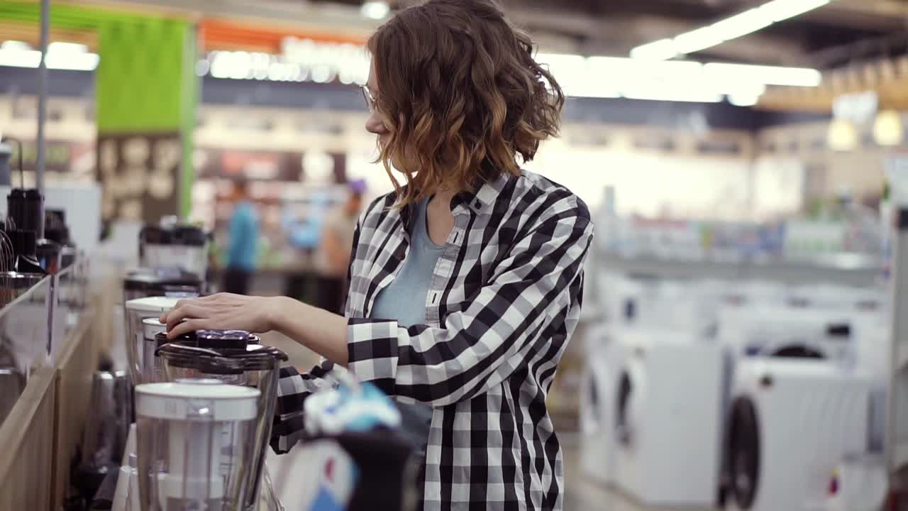 en la tienda de electrodomésticos, una mujer morena rizada en una camisa a cuadros elige una licuadora para comprar viendo y sosteniendo el