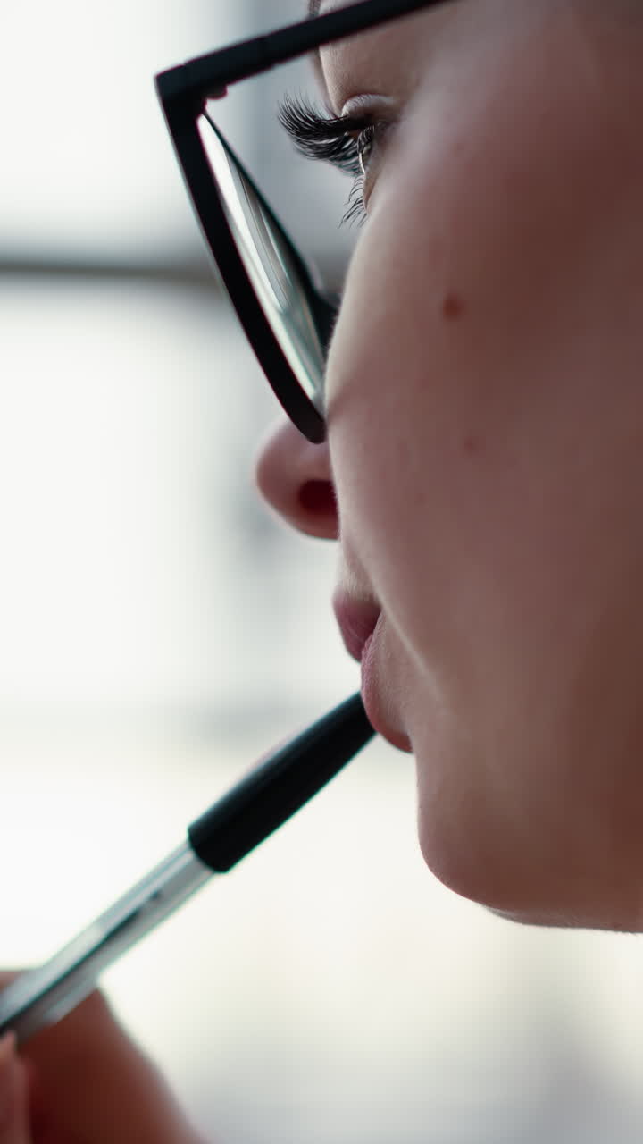 vista cercana de la cara de la dama con gafas y pluma en los labios reflejando algo, perdida en el pensamiento con un fondo suave y borroso, mostrando un momento reflexivo y reflexivo