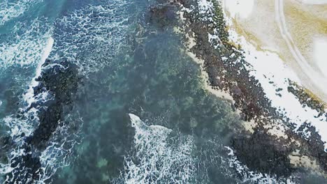 Toma-Aérea-Apuntando-Hacia-Sandgerdi,-Costa-De-Islandia-Que-Muestra-Olas-Blancas-Y-Agua-Clara-Con-Rocas-Negras