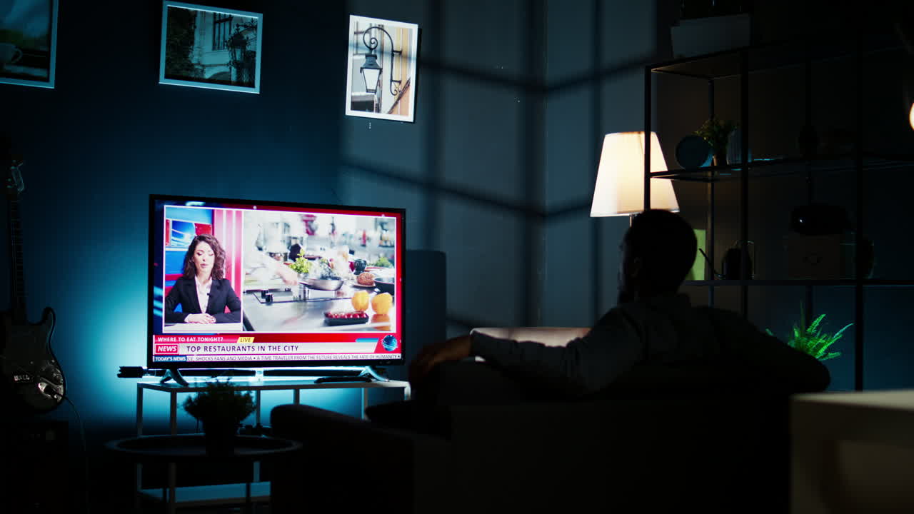 젊은 남자는 거실의 소파에서 휴식을 취하고 tv에서 최신 헤드라인을 따르고 있습니다.