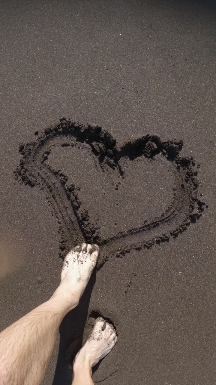 el pie masculino dibuja un corazón en la arena de la playa y una ola lo lava.