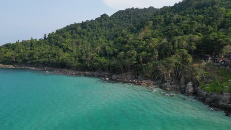 Aerial-view-of-coastal-landscape-featuring-crystal-clear-turquoise-water-along-the-rocky-shore-embraced-by-a-lush,-green-tropical-forest-under-a-clear-sky