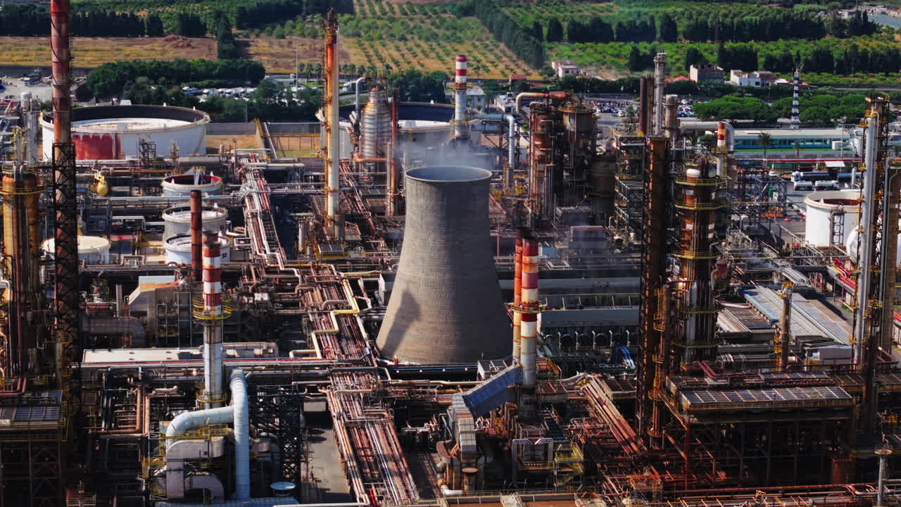 Drone panning shot over an oil refinery chimney in Sicily, with light smoke and industrial structures around