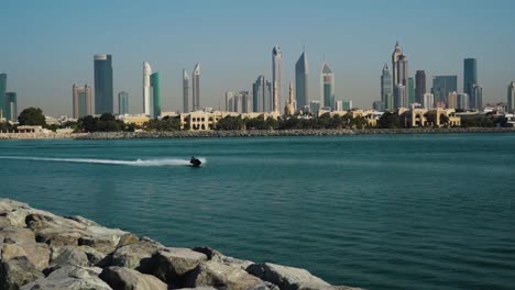 Man-Jet-Skiing-At-Persian-Gulf-Leaving-Wake-On-Surface-With-Dubai-Skyline-And-Jumeirah-1-In-The-Background-From-Pearl-Jumeirah,-Dubai,-UAE