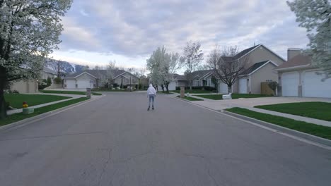 Un-Hombre-Desde-Atrás-Patinando-Por-Una-Subdivisión-Del-Vecindario-Con-Montañas-Cubiertas-De-Nieve-En-El-Fondo-En-Utah